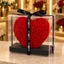 Coração vermelho de rosas de espuma (15 cm) em caixa transparente com fita preta ENROSED, sobre um balcão bege no showroom.
